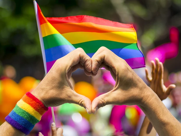 LGBTQ+-Herz Hände formen ein Herz vor einer Regenbogenflagge