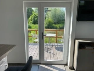 Blick aus einem hellen Raum auf eine Holzterrasse mit Tisch und Stühlen vor einem kleinen Teich.