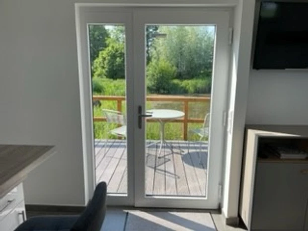 Tiny-Teich-Haus_Beatrix_Boekstegers_1.jpg Blick aus einem hellen Raum auf eine Holzterrasse mit Tisch und Stühlen vor einem kleinen Teich.