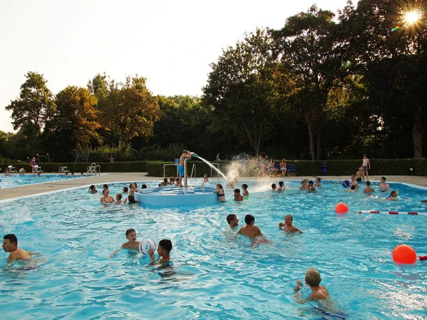 Sommerschwimmbad mit vielen Menschen im Wasser, umgeben von Bäumen, während die Sonne scheint.