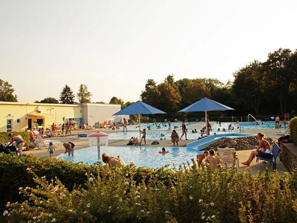 Außenansicht eines lebhaften Freibads mit mehreren Schwimmbecken, Sonnenliegen und Besuchern unter Schirmen.