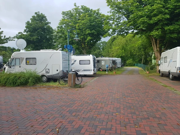 Wohnmobilstellplatz Seglertreff Otterndorf Weiße Wohnmobile parken auf einem gepflasterten Platz unter grünen Bäumen in Otterndorf.