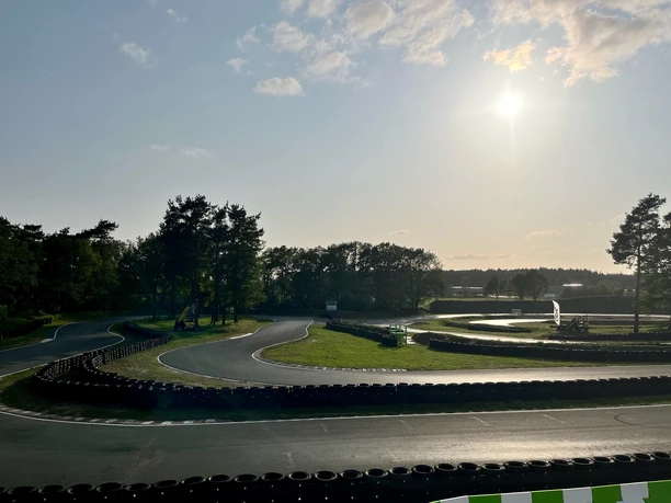 Sonnenbeschienene Outdoor-Kartbahn mit geschwungenem Asphaltkurs, umgeben von Bäumen und Reifenbarrieren.