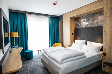 Modern eingerichtetes Hotelzimmer mit Doppelbett, türkisfarbenen Vorhängen und Holzakzenten.