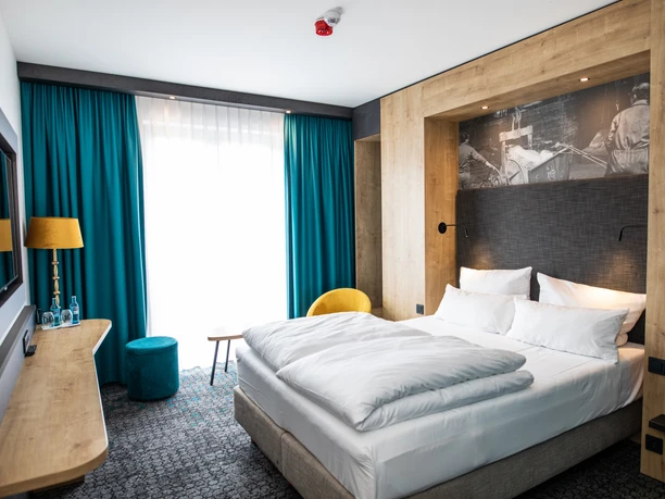 0167.jpg Modern eingerichtetes Hotelzimmer mit Doppelbett, türkisfarbenen Vorhängen und Holzakzenten.