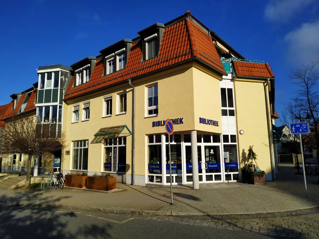Stadtbibliothek Neustadt in Sachsen Gelbes Gebäude mit rotem Dach, beschriftet mit "Bibliothek", bei sonnigem Wetter; Fahrradständer und Verkehrsschilder im Vordergrund.