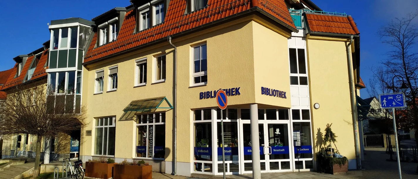 Stadtbibliothek Neustadt in Sachsen Gelbes Gebäude mit rotem Dach, beschriftet mit "Bibliothek", bei sonnigem Wetter; Fahrradständer und Verkehrsschilder im Vordergrund.
