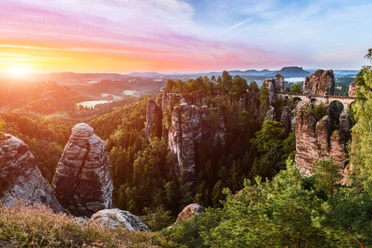 Rock formations and a stone bridge in a green forest landscape at sunset, with a colorful sky in the background.