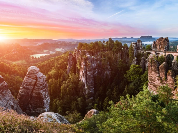 Blick von der Ferdinandsaussicht auf die Bastei Felsformationen und eine Steinbrücke in einer grünen Waldlandschaft bei Sonnenuntergang, mit einem farbenfrohen Himmel im Hintergrund.