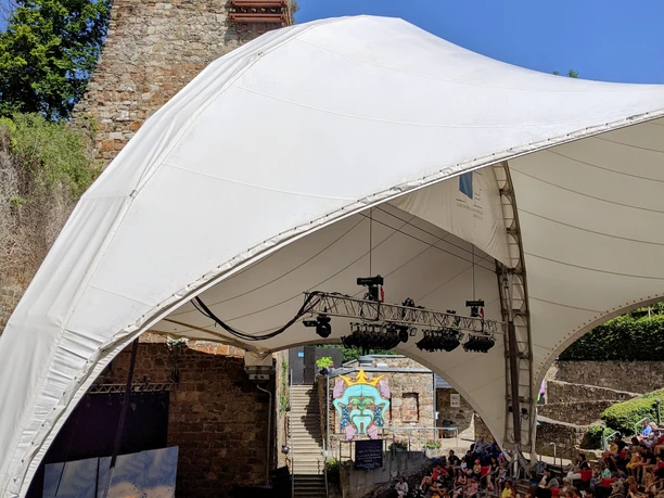 DDT_BurgWilhelmstein_StimmungPanama(C)DDT.jpg Unter einem großen Zeltdach wird ein Theaterstück auf einer Bühne aufgeführt, um die viele Zuschauer auf einer halbkreisförmigen Tribüne sitzen.