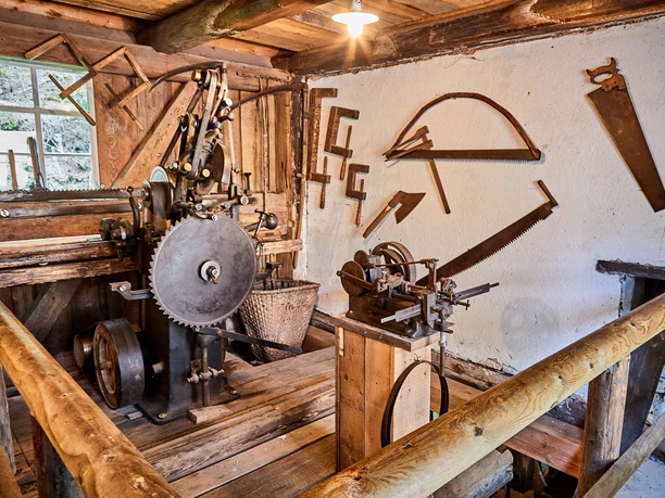 Austellung Neumannmühle Alte Werkstatt mit Holzsägemaschine, umgeben von hölzernen Wänden und Werkzeugen an der Wand; rustikale, historische Atmosphäre.