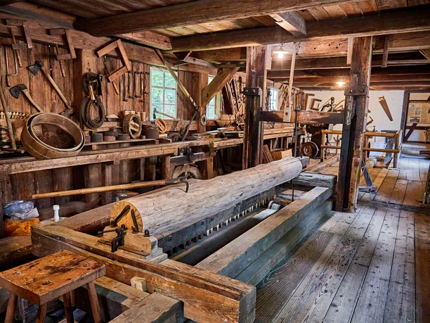 Austellung Neumannmühle Alte Holzwerkstatt mit rustikalen Werkzeugen und Holzbalken, ein großer Baumstamm liegt auf einer historischen Säge, warme, nostalgische Atmosphäre.