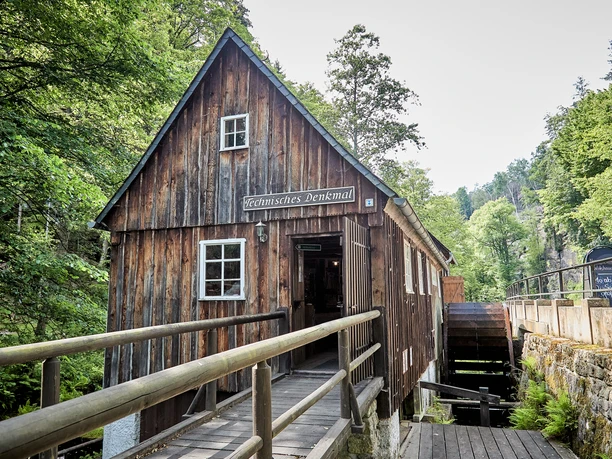 Wooden building with "Technical Monument" sign, surrounded by trees, with a water wheel on the side and a wooden footbridge in the foreground.