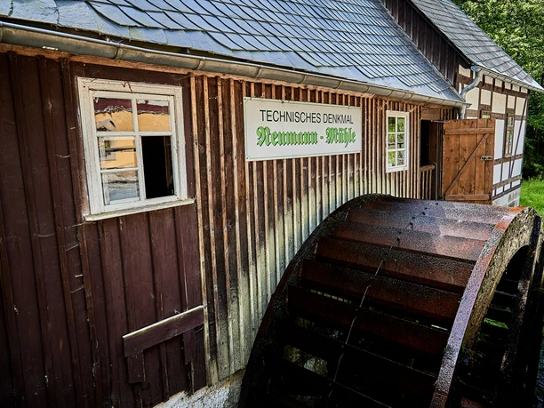 Historic watermill with wooden wheel, brown façade and a sign "Technisches Denkmal Neumann-Mühle" in green lettering.