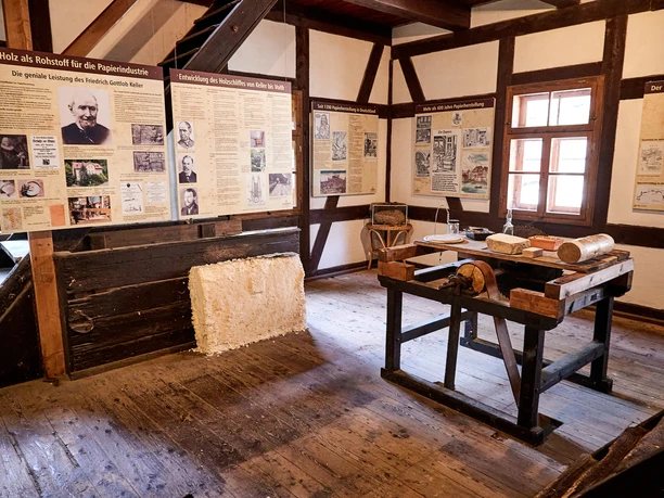 A historic room with a wooden floor houses an exhibition on paper production, with information boards, a wooden table and tools.