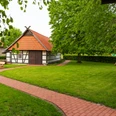 Hille Holzhausen II - Alte Scheune und Historische Dorfschmiede Fachwerkgebäude mit roten Ziegeldächern, umgeben von grüner Wiese und Bäumen, ein Fußweg führt entlang.