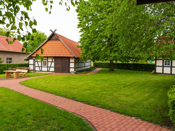 Hille Holzhausen II - Alte Scheune und Historische Dorfschmiede Fachwerkgebäude mit roten Ziegeldächern, umgeben von grüner Wiese und Bäumen, ein Fußweg führt entlang.