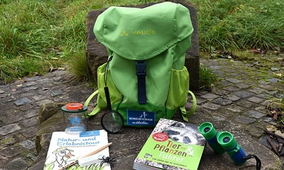 Entdeckerrucksack am Naturerlebnispfad in Monheim am Rhein Ein grüner Rucksack mit Büchern, einer Lupe und einem Fernglas liegt auf einem Naturpfad.