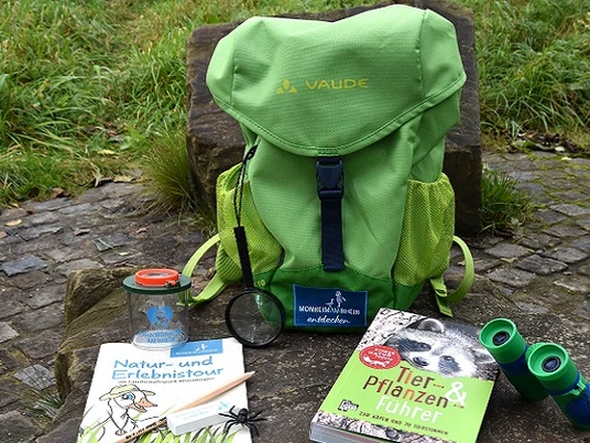 Entdeckerrucksack am Naturerlebnispfad in Monheim am Rhein Ein grüner Rucksack mit Büchern, einer Lupe und einem Fernglas liegt auf einem Naturpfad.