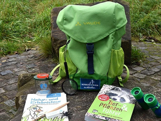 Entdeckerrucksack am Naturerlebnispfad in Monheim am Rhein Ein grüner Rucksack mit Büchern, einer Lupe und einem Fernglas liegt auf einem Naturpfad.