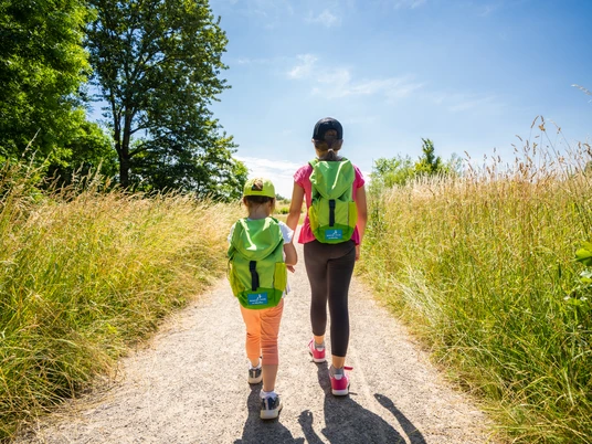 Mit dem Entdeckerrucksack auf dem Naturerlebnispfad in Monheim am Rhein Zwei Personen mit Rucksäcken spazieren auf einem sonnigen Pfad durch hohe Gräser.