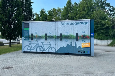 Fietsgarage met genummerde vakken en blauw muurontwerp, omringd door bomen en geplaveide grond, onder een heldere hemel.