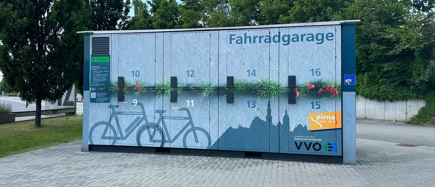 Fietsgarage met genummerde vakken en blauw muurontwerp, omringd door bomen en geplaveide grond, onder een heldere hemel.