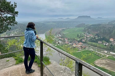 Aussicht genießen auf dem Kanapee Frau in Jeansjacke steht an einem Geländer und blickt auf eine grüne Landschaft mit Fluss und Dorf, umgeben von Hügeln unter bewölktem Himmel.