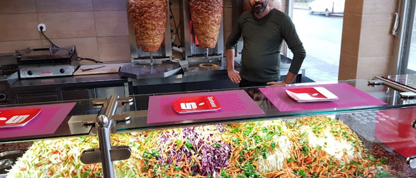 Ein Mann steht hinter einer Theke, auf der bunte Gemüsearten in Reihen angeordnet sind. Im Hintergrund drehen sich zwei Döner-Spieße.