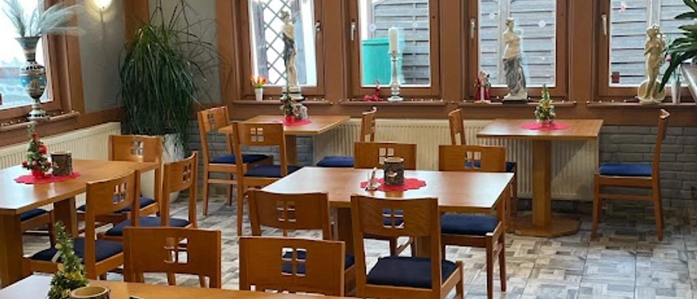 Restaurant mit Holzmöbeln, dekorativen Pflanzen und großen Fenstern, die viel Tageslicht einlassen.