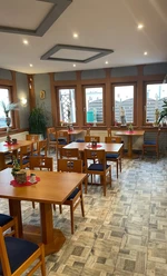 Greek House Grill Restaurant mit Holzmöbeln, dekorativen Pflanzen und großen Fenstern, die viel Tageslicht einlassen.