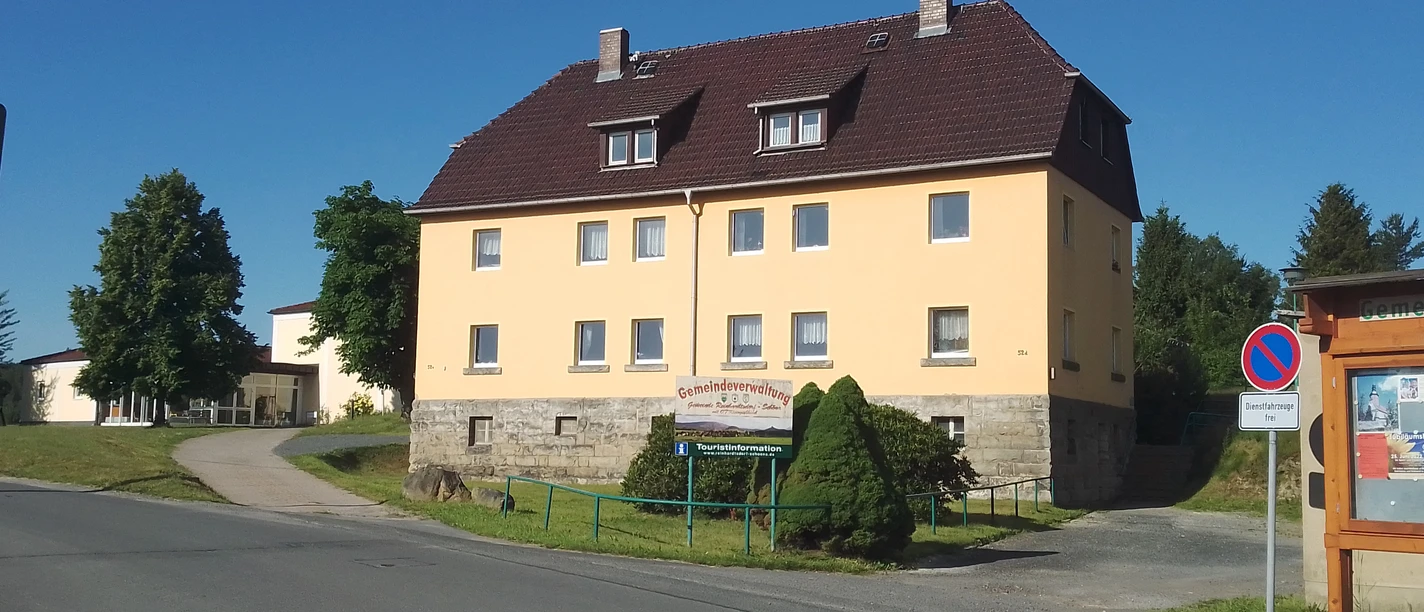 Yellow two-story building with a brown roof, surrounded by trees, under a clear sky; a sign indicates the municipal administration.