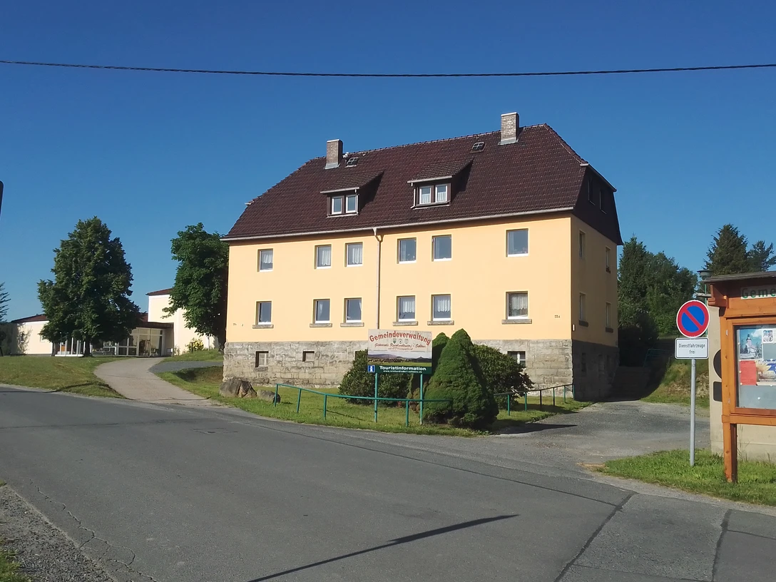 Touristinformation Reinhardtsdorf-Schöna Gelbes zweistöckiges Gebäude mit braunem Dach, umgeben von Bäumen, bei klarem Himmel; ein Schild weist auf die Gemeindeverwaltung hin.