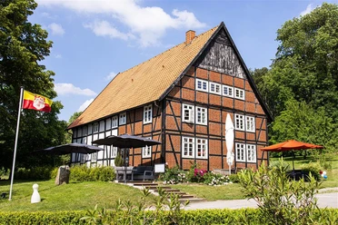 Fachwerkhaus mit roten Ziegeln und großem Satteldach, umgeben von grüner Landschaft und Sonnenschirmen.