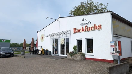 Eine weiße Gebäudefront mit der Aufschrift "Wester's Backfuchs", Schilder und ein Terrassenbereich.