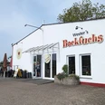 Backfuchs Eine weiße Gebäudefront mit der Aufschrift "Wester's Backfuchs", Schilder und ein Terrassenbereich.