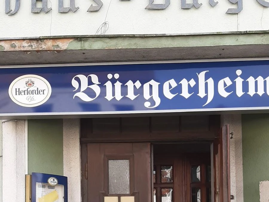 speisekartenweb.jpg Eingang des Restaurants Bürgerheim mit blauem Schild und Herforder-Logo, Holztür und Fenster.