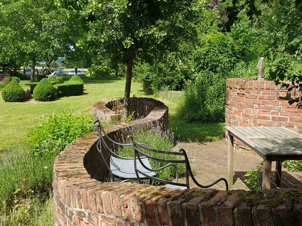 Grüne Gartenlandschaft mit geschwungener Backsteinmauer, hölzernem Tisch, Bänken und buschigem Grün.