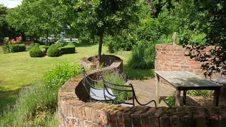 Garten Wasels Grüne Gartenlandschaft mit geschwungener Backsteinmauer, hölzernem Tisch, Bänken und buschigem Grün.