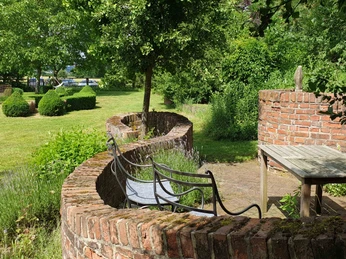 Garten Wasels Grüne Gartenlandschaft mit geschwungener Backsteinmauer, hölzernem Tisch, Bänken und buschigem Grün.