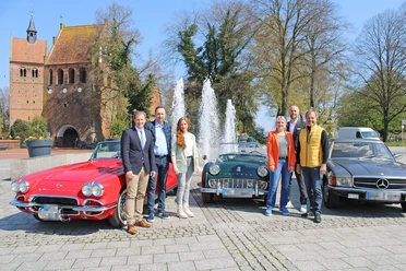 Classic Car Charity Bad Zwischenahn.jpg Eine Gruppe Menschen posiert vor drei klassischen Autos, darunter ein rotes Cabriolet, auf einem Platz mit Springbrunnen im Hintergrund.