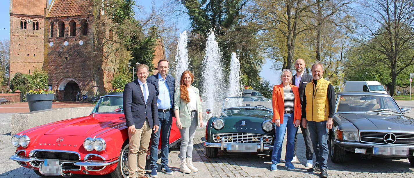 Classic Car Charity Bad Zwischenahn.jpg Eine Gruppe Menschen posiert vor drei klassischen Autos, darunter ein rotes Cabriolet, auf einem Platz mit Springbrunnen im Hintergrund.