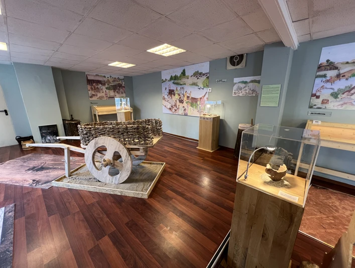 Ausstellungsraum mit historischen Werkzeugen und Infotafeln auf Holzboden, Fokus auf großen Holzwagen.