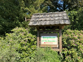 Ein Holzschild mit der Aufschrift "Spargelessen auf dem Bauernhof", umgeben von grünen Büschen.
