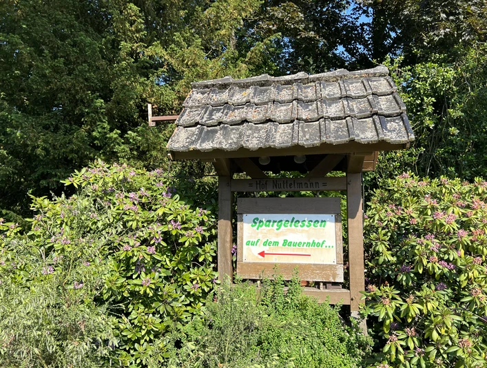 Spargelhof Nuttelmann Ein Holzschild mit der Aufschrift "Spargelessen auf dem Bauernhof", umgeben von grünen Büschen.