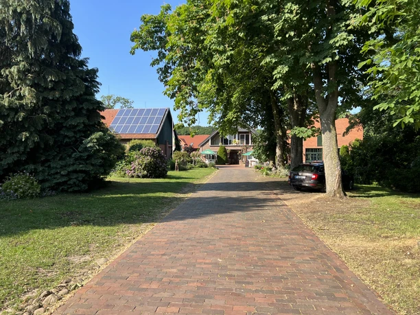 Roter Backsteinweg führt zu einem Hof mit Solardach zwischen grünen Bäumen und Sträuchern.