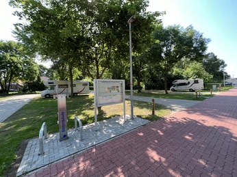 Wohnmobilstellplatz mit Stromsäule, Infotafel, zwei Wohnmobile, von Bäumen gesäumt bei klarem Himmel.