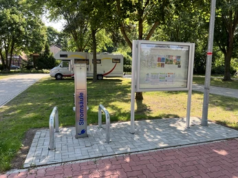 Wohnmobilstellplatz mit Stromsäule und Infotafel, umgeben von Bäumen und einem ruhigen Umfeld.