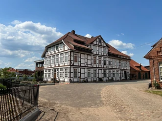 Fachwerkhaus mit rotem Dach, umgeben von gepflastertem Hof unter blauem Himmel mit Wolken.