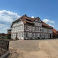 Fachwerkhaus mit rotem Dach, umgeben von gepflastertem Hof unter blauem Himmel mit Wolken.
